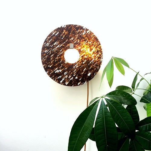 Applique murale en plastique recyclé de couleur marron foncé tachetée de blanc et de forme circulaire. L'applique est accrochée au mur au-dessus d'une plante verte et est allumée.