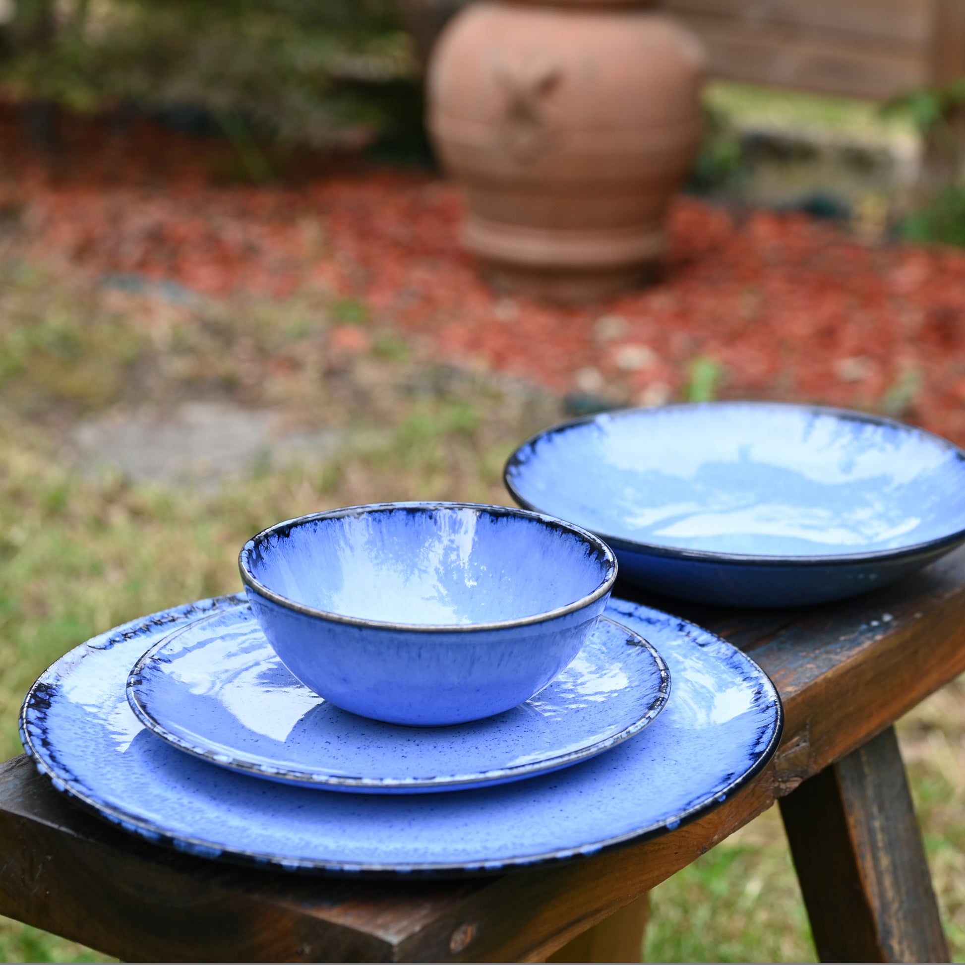 Assiettes de différentes tailles (plate, à dessert, creuse) et bol en grès naturel avec émail réactif bleu profond, empilées sur un banc en bois dans un jardin