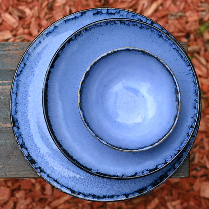 Assiettes de différentes tailles (plate, à dessert, creuse) et bol en grès naturel avec émail réactif bleu profond, empilées sur un banc en bois dans un jardin