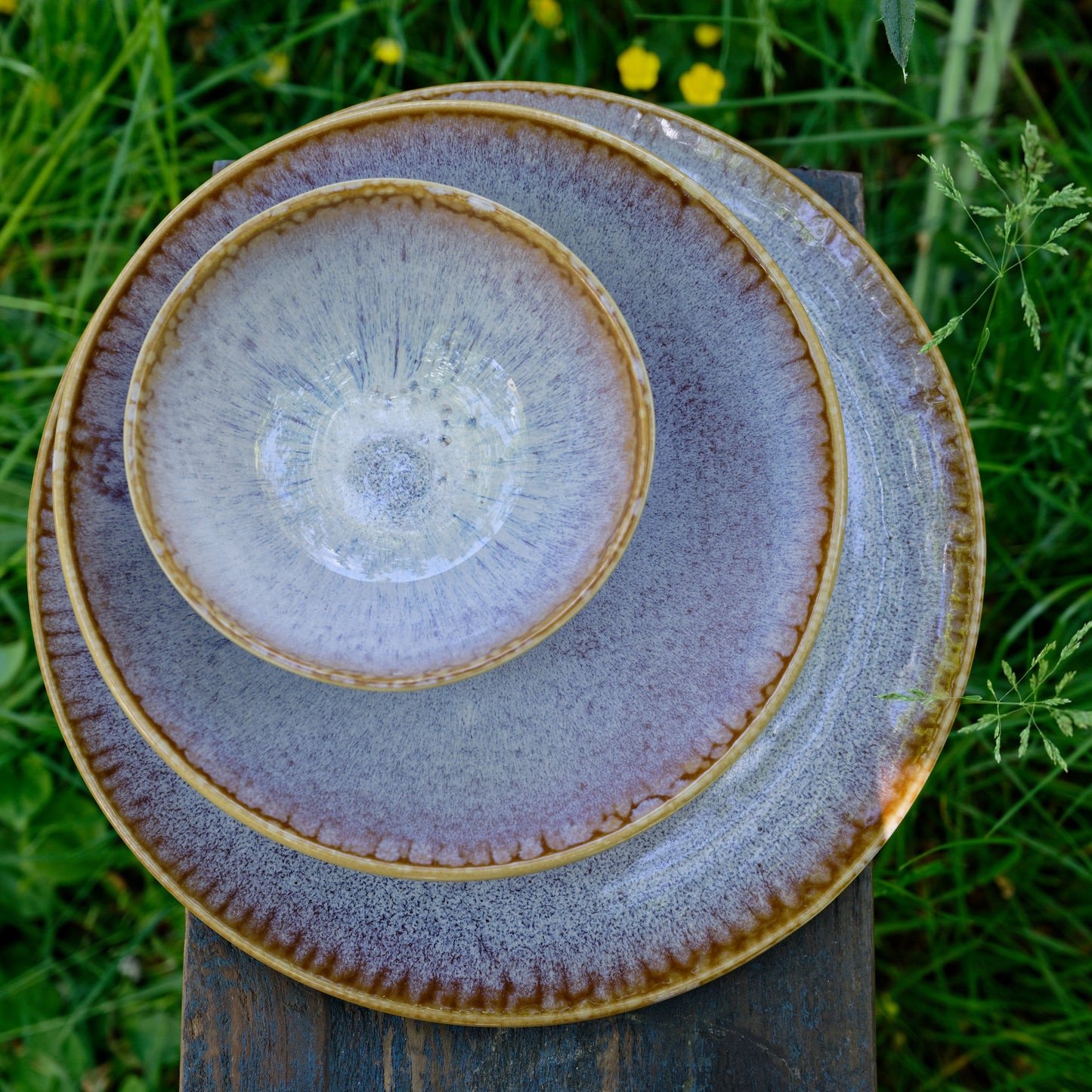 Assiettes de différentes tailles (plate, à dessert, creuse) et bol en grès naturel avec émail réactif ambre et violet subtil, empilées sur un banc en bois dans un jardin