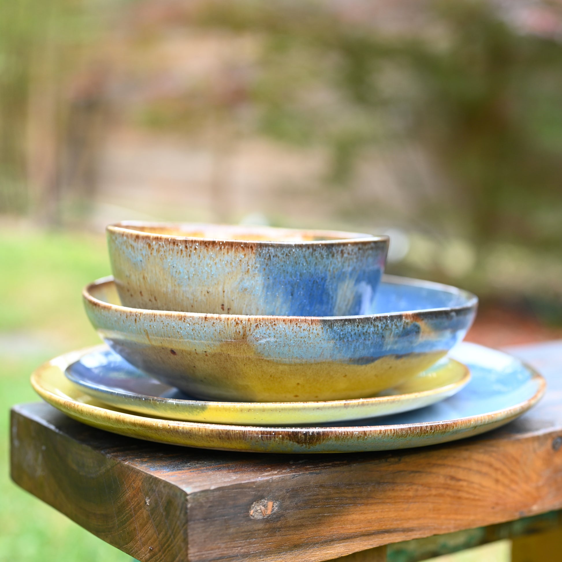 assiettes de différentes tailles (plates, à dessert, creuse) et bol de style "Corals" (grès naturel avec motif tricolore bleu profond, bleu ciel et jaune, empilées sur un banc en bois dans un jardin