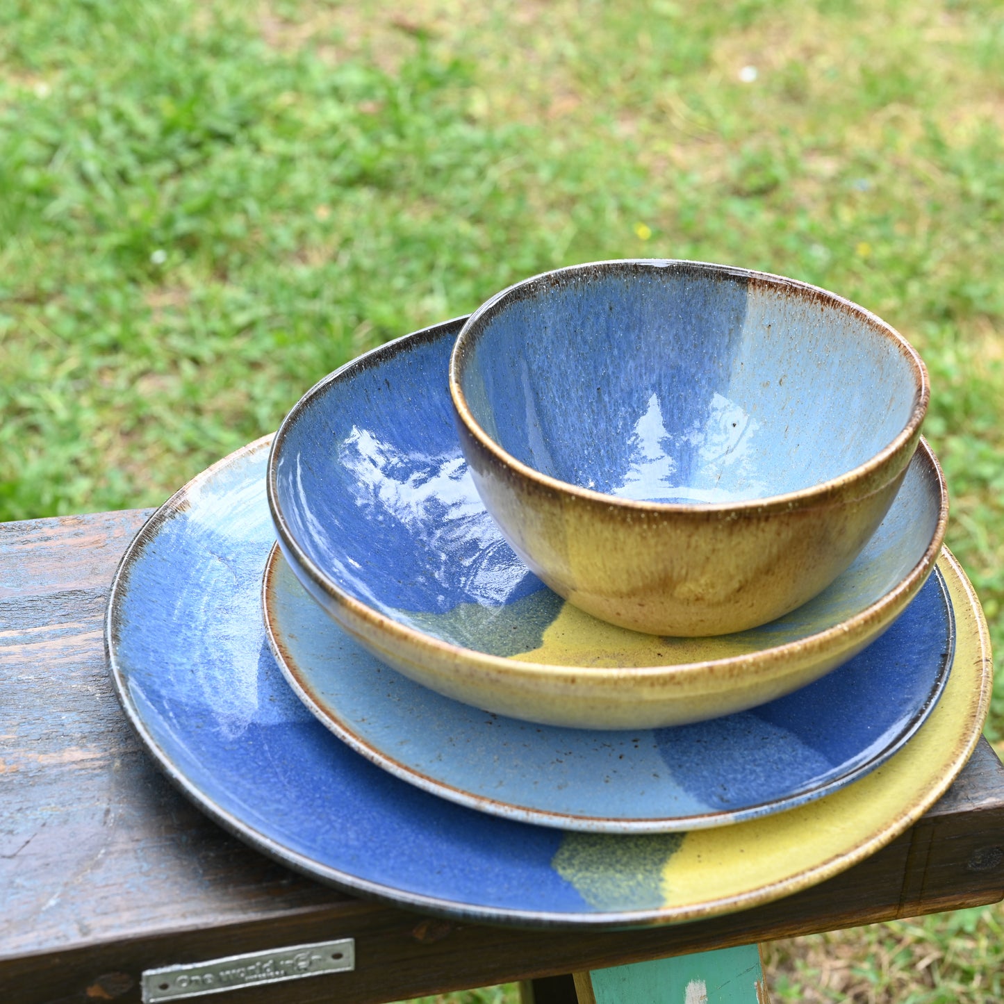 assiettes de différentes tailles (plates, à dessert, creuse) et bol de style "Corals" (grès naturel avec motif tricolore bleu profond, bleu ciel et jaune, empilées sur un banc en bois dans un jardin