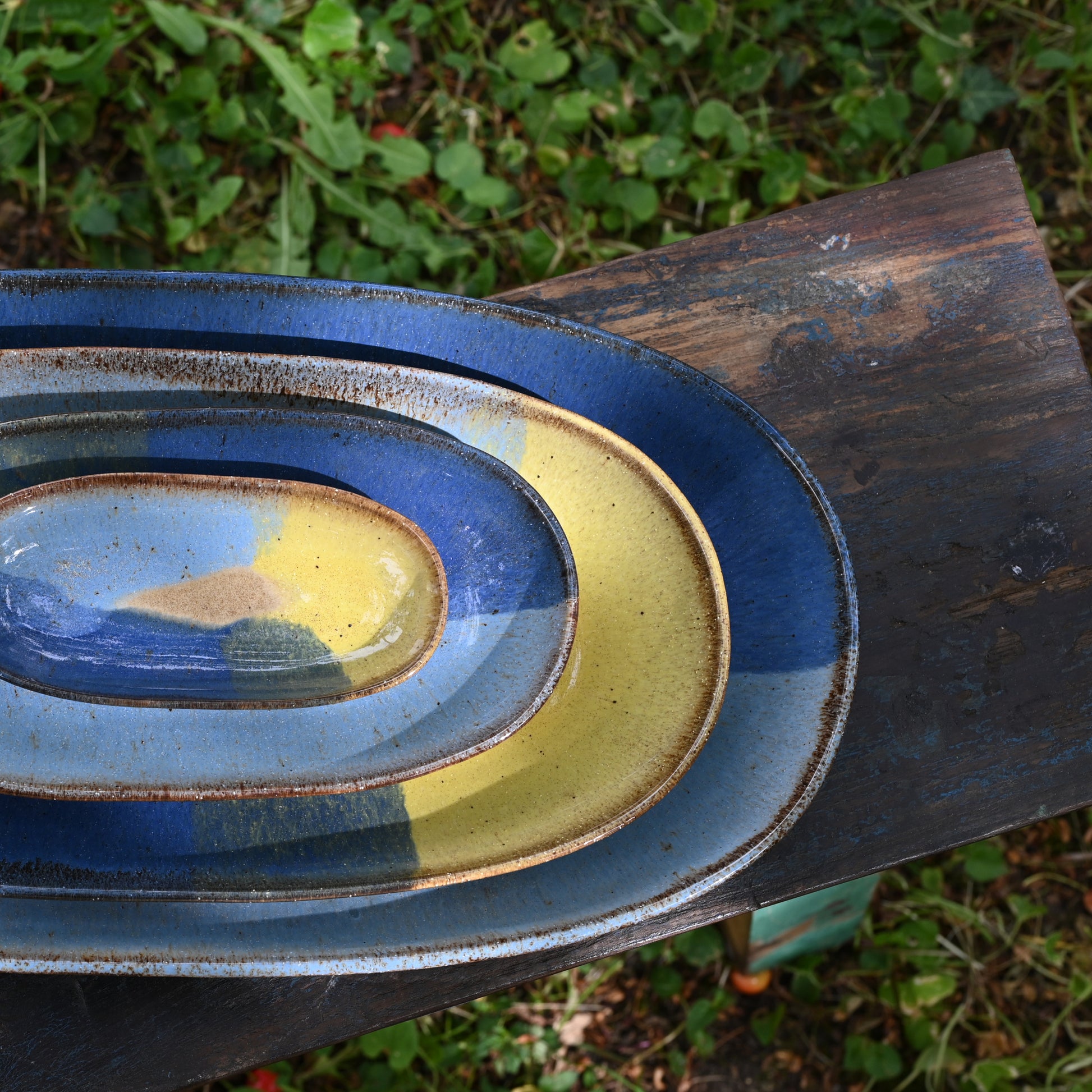 vue de dessus, 4 plats à service en grès naturel avec motif tricolore bleu profond, bleu ciel et jaune, empilés sur un banc dans un jardin