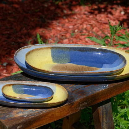 vue de biais, plats à service en grès naturel avec motif tricolore bleu profond, bleu ciel et jaune, sur un banc dans un jardin